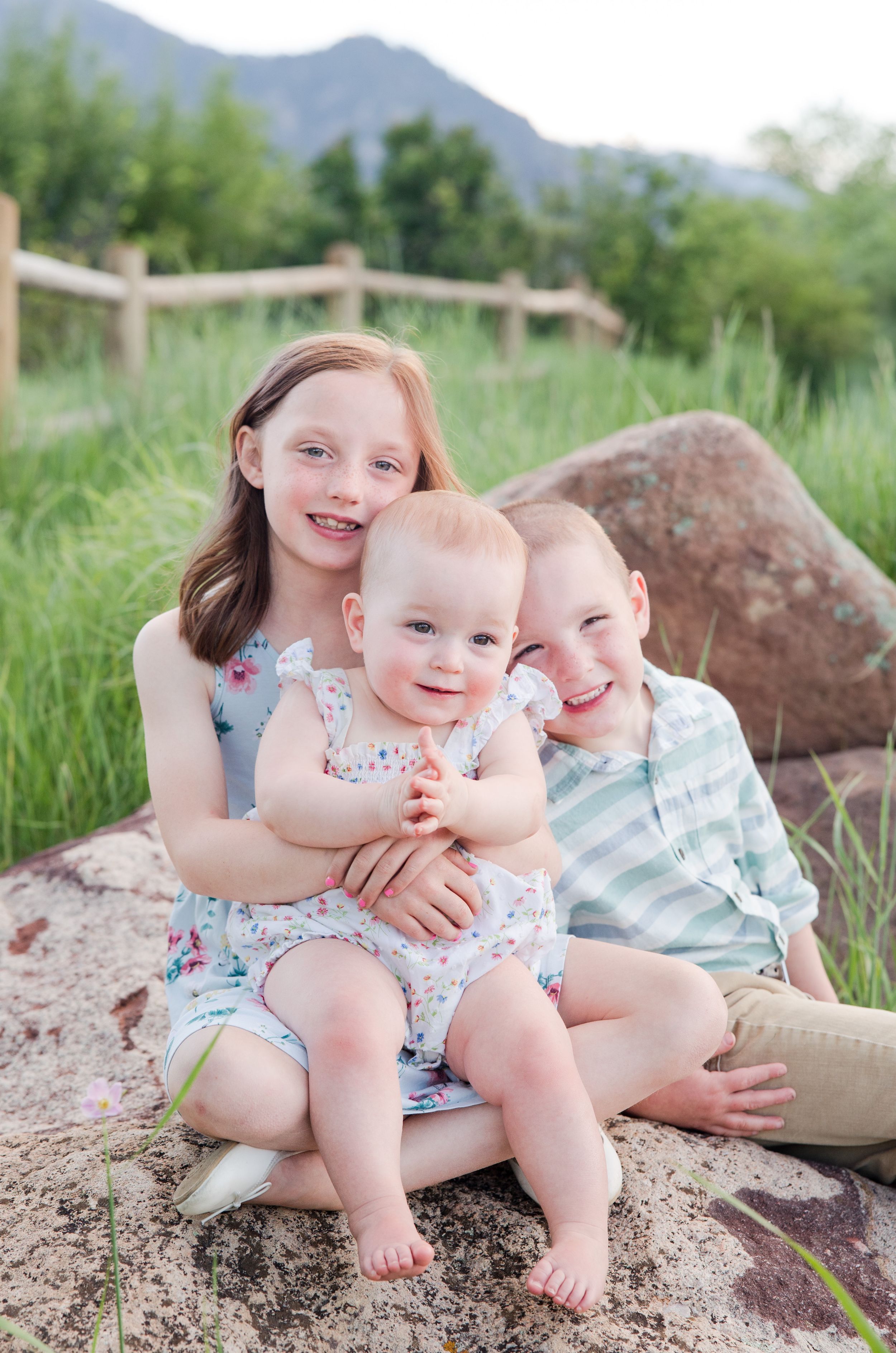 Three candid photos of siblings sitting together on rocks against a mountain backdrop show loving family moments in nature.