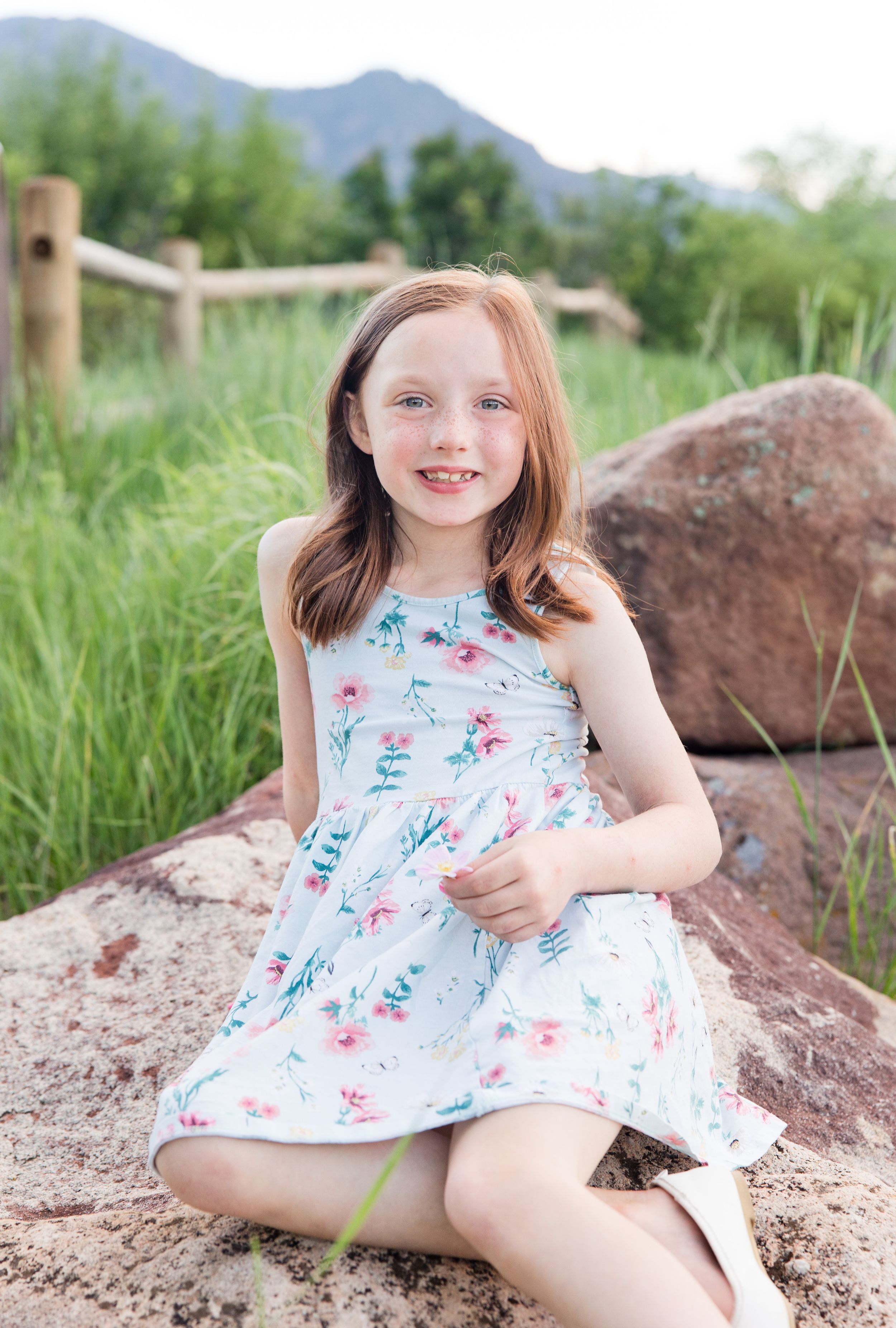 Young person in a floral summer dress sits on red rocks with mountains and greenery in the background.