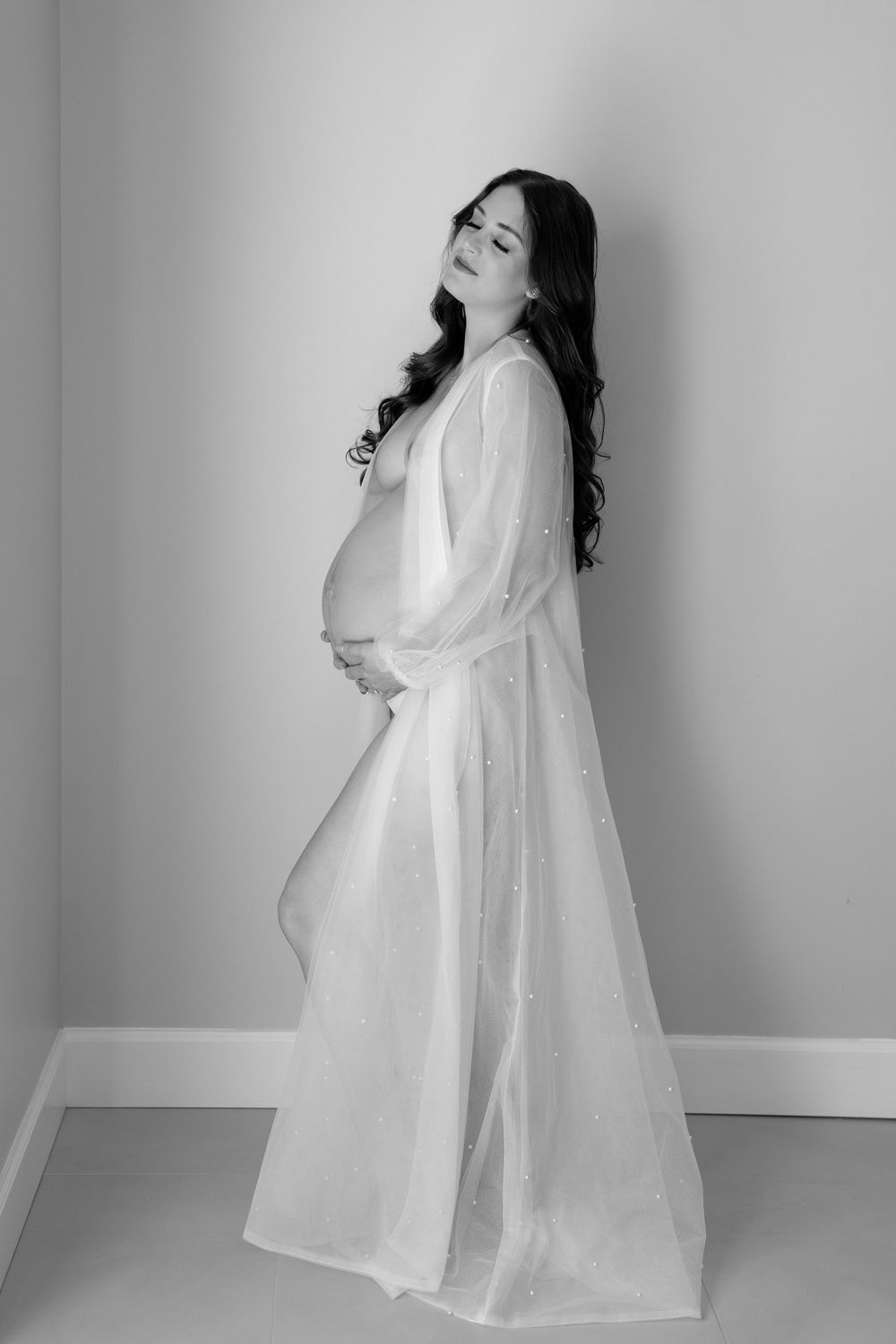 Artistic black and white maternity photo featuring an expectant mother in a flowing white dress against a simple backdrop.