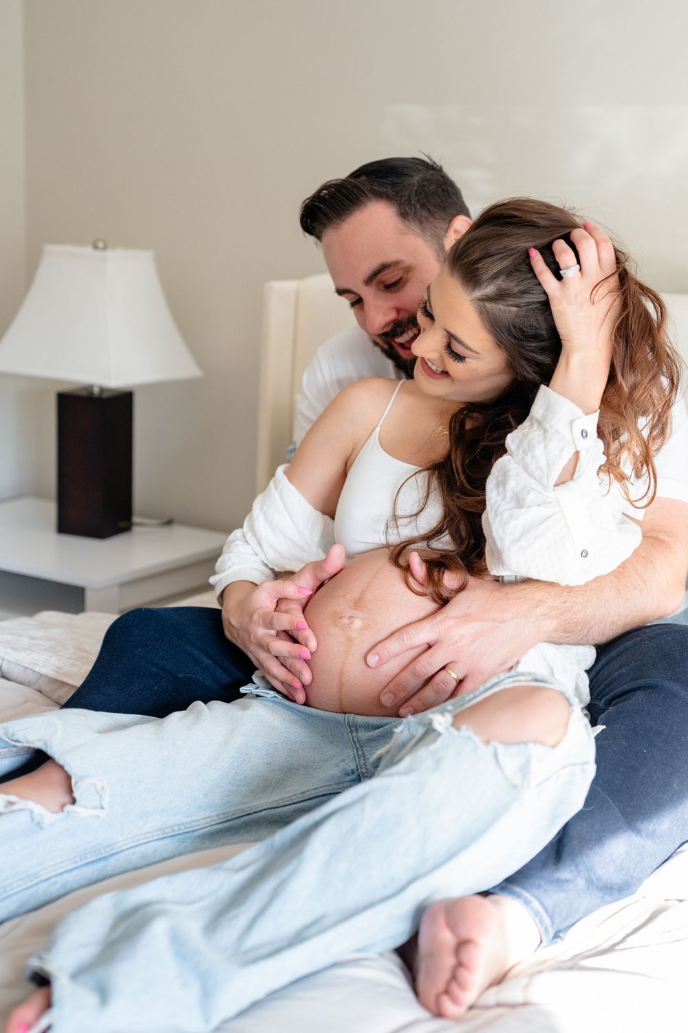 Intimate maternity moment captured on a white bed with soft natural lighting.