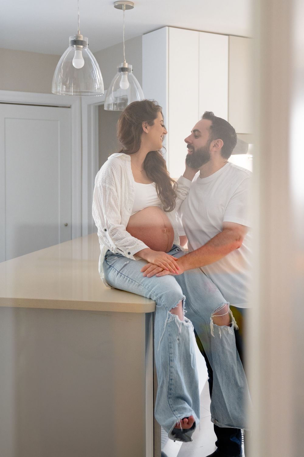 Expecting couple shares an intimate moment in bright modern kitchen with pendant lights.