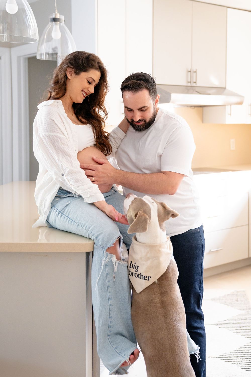 Couple shares intimate moment in bright modern kitchen with their dog joining in celebration of pregnancy.