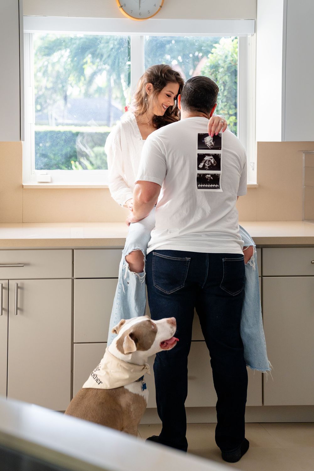 Couple embraces in kitchen with dog looking up at them near window with ultrasound photos.