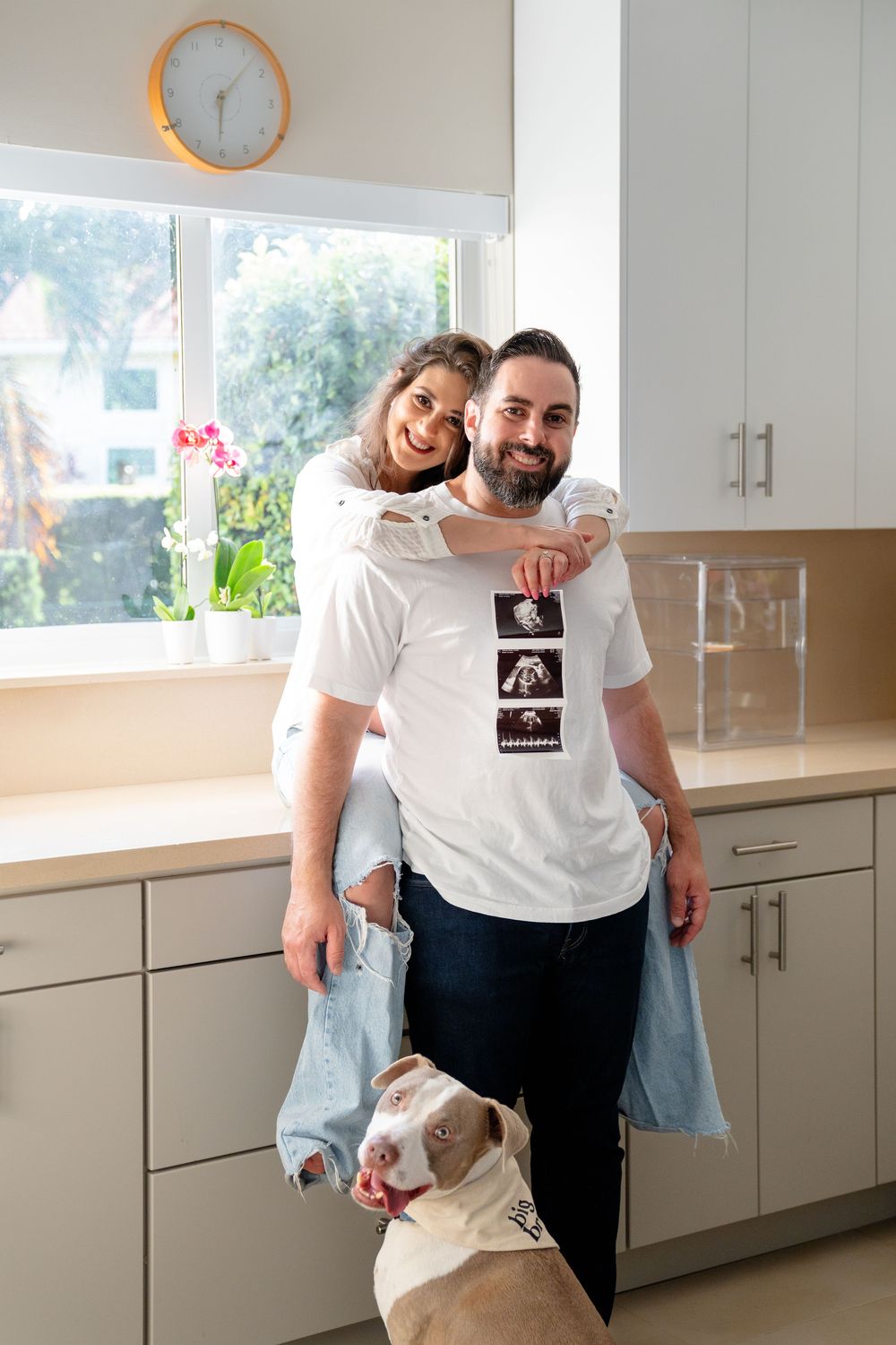 A couple poses in their bright kitchen with their pit bull dog while wearing casual clothes and smiling.