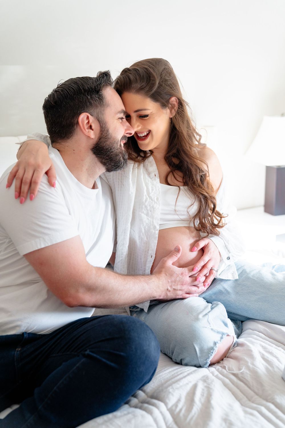 A couple shares tender moments while relaxing together on white bedding in a bright, airy setting.