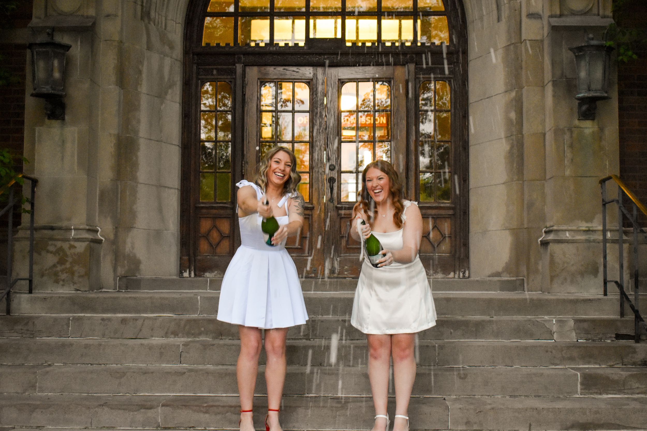 Two bridesmaids in white dresses hold bouquets while standing on stone steps in front of ornate church entrance.
