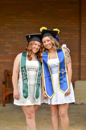 Two graduates in white dresses wearing ceremonial stoles and mortarboard caps pose together against a brick wall.