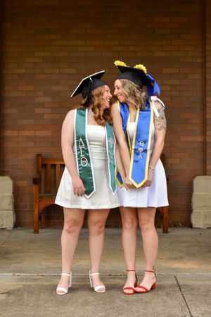 Two graduates in white dresses and graduation caps wearing academic stoles share a special moment against a brick wall.