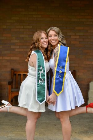 Two sorority members wearing white dresses and graduation stoles pose together against a brick wall.