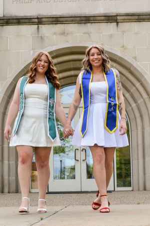 Two graduates in white dresses wearing academic stoles and sashes pose together outside a stone building entrance.