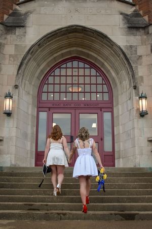 Two people in light dresses walk up stone steps toward a large arched church entrance with burgundy doors.
