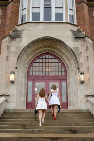 Two people in white dresses climb stone steps toward an ornate arched entrance with red doors of a brick building.