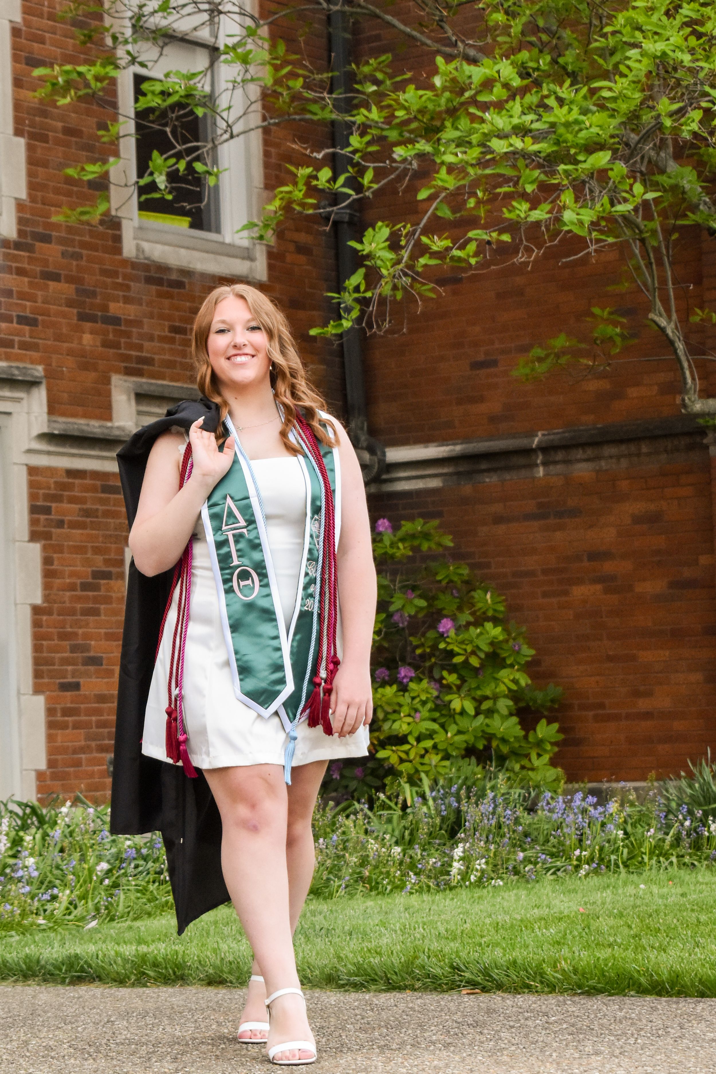 Graduate in white dress and green sash walking confidently on campus path with red brick building and greenery in background.