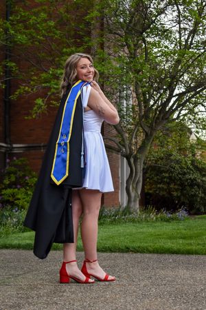 Graduate in white dress and graduation stole with blue and gold honors cord poses outdoors on campus grounds.