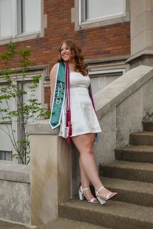Graduate in a white dress and green graduation stole poses on concrete steps of brick building.