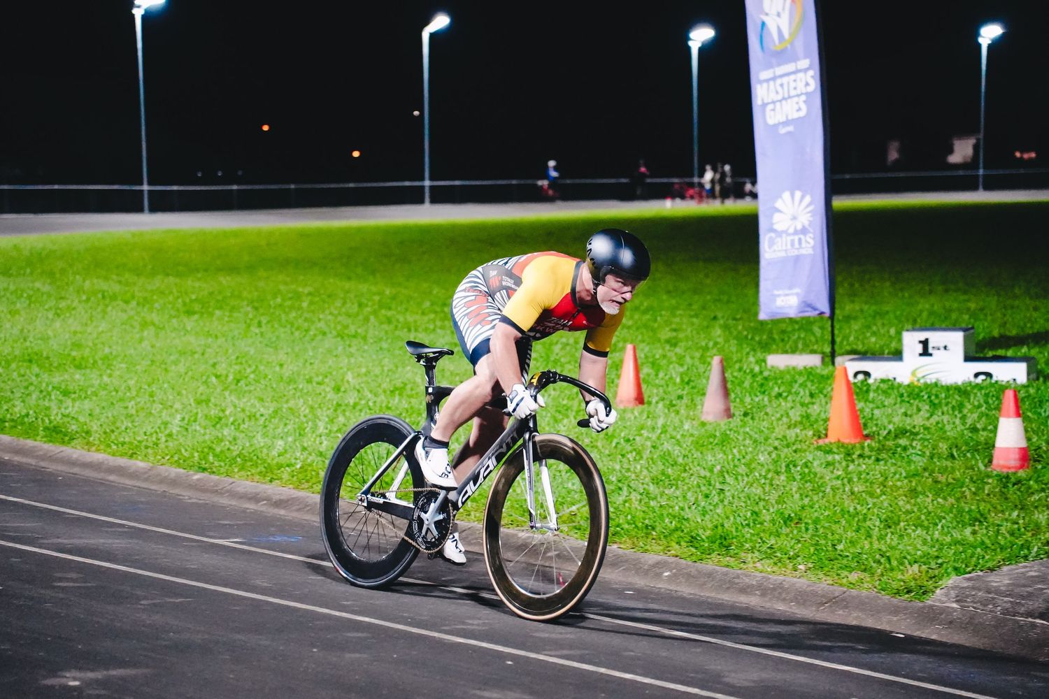 A cyclist in racing gear leans forward on their bike while training on an illuminated track at night.