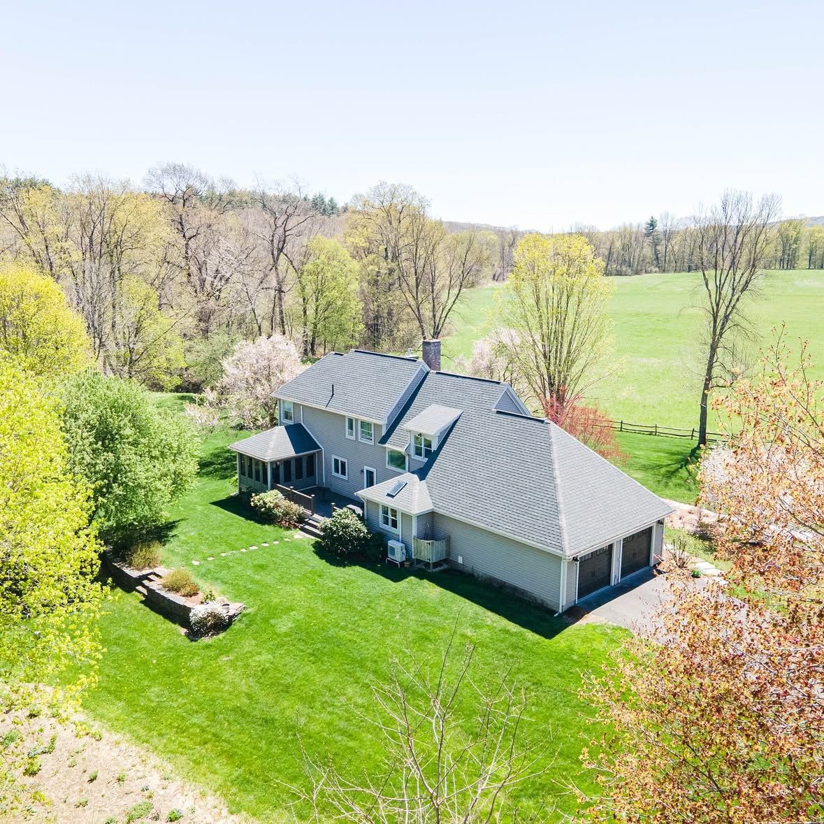 Aerial view of a gray ranch home with attached garage situated on a spacious green lawn surrounded by spring trees.