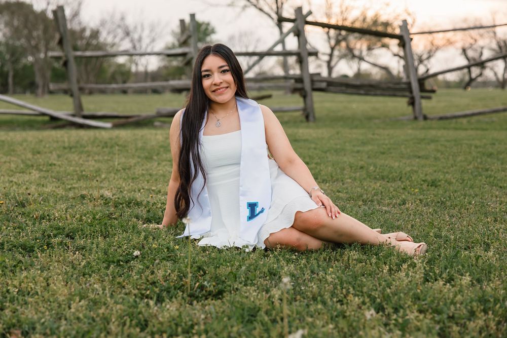 A woman in a flowing white sundress sits gracefully on grass in a park during golden hour.