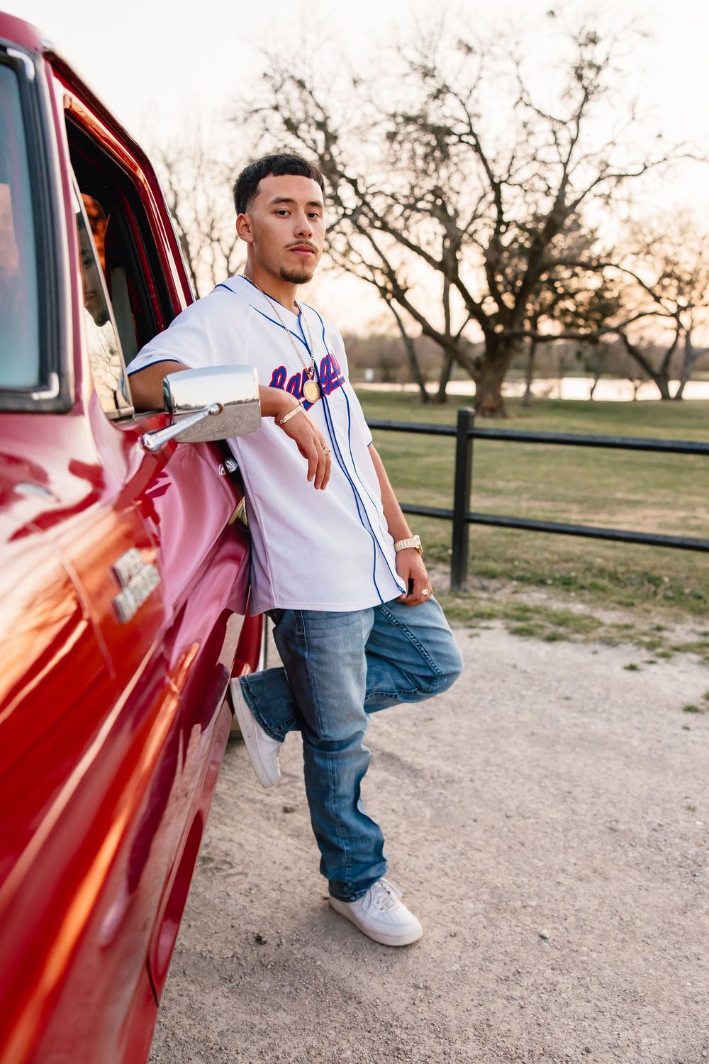 Person in white baseball jersey and jeans leans against vintage red car in park setting during sunset.