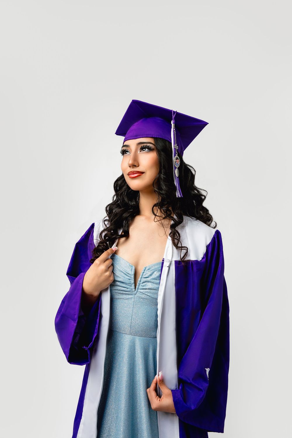 Graduate in purple cap and gown over light blue dress poses against white background.