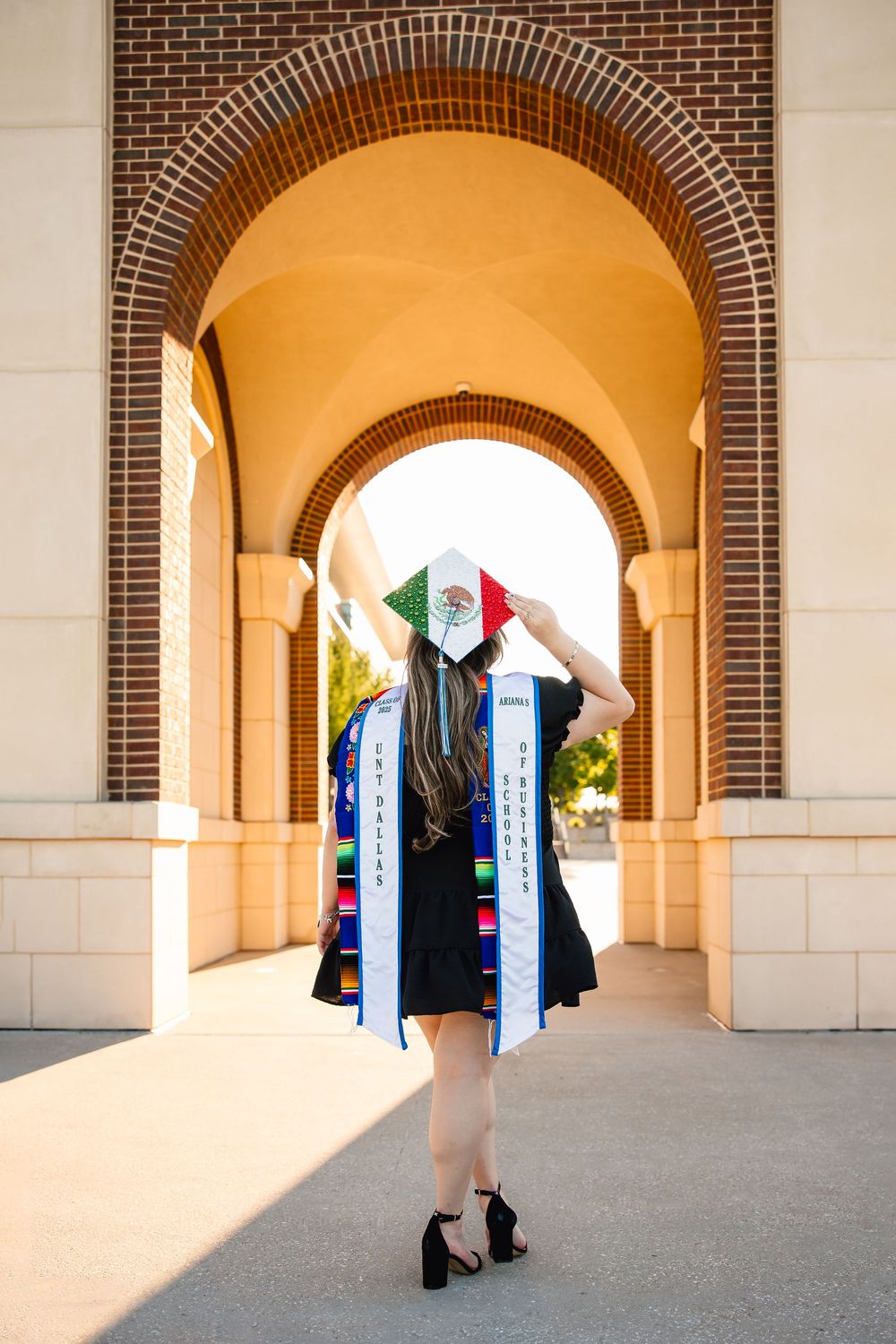 Graduate wearing decorated graduation stole and cap stands in arched brick doorway showing Mexican flag colors.