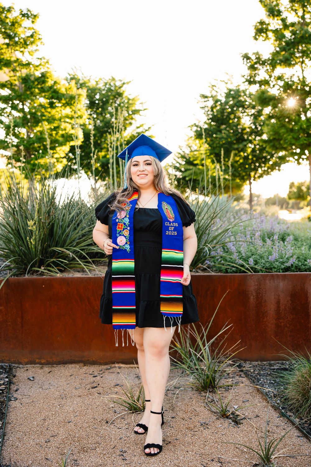Graduate wearing blue cap, black dress and colorful graduation stole stands in outdoor garden setting at sunset.