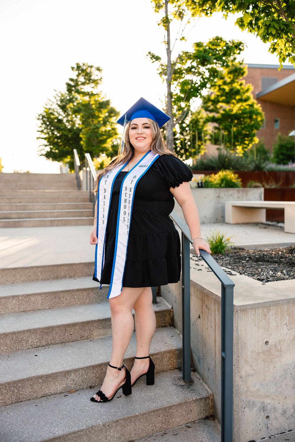 Graduate in blue cap and black dress stands on concrete steps during sunset with trees in background.