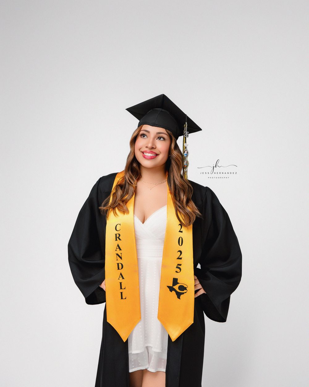 Graduate in a black cap and gown with gold stole smiles while posing against gray background for graduation photo.