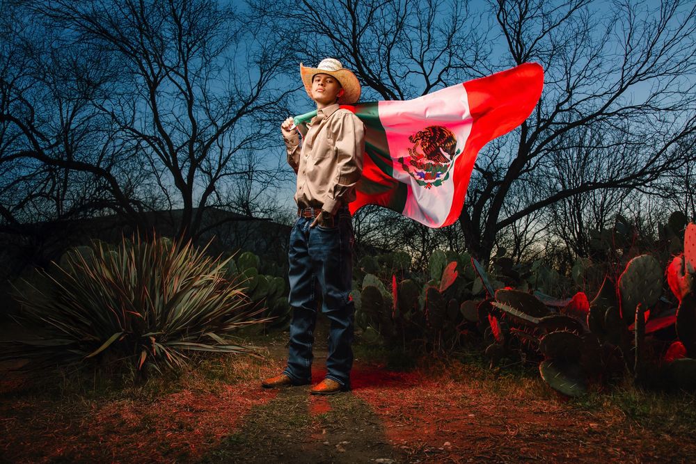 A person stands holding a Canadian flag at dusk against a backdrop of bare trees and desert plants.