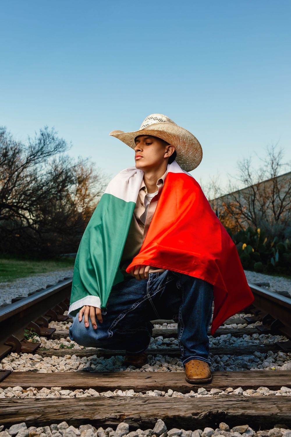 A figure in a cowboy hat and colorful clothing sits contemplatively on railroad tracks against a bright blue sky.