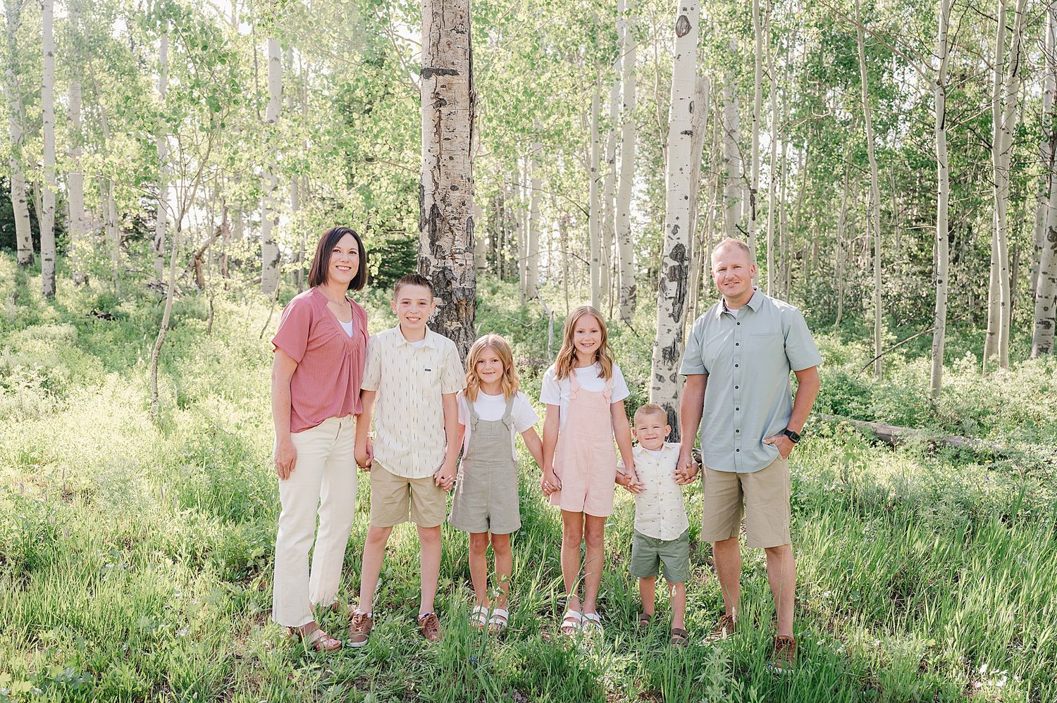 Family wearing pink and blue outfits poses together in sun-dappled aspen forest during summer photoshoot.