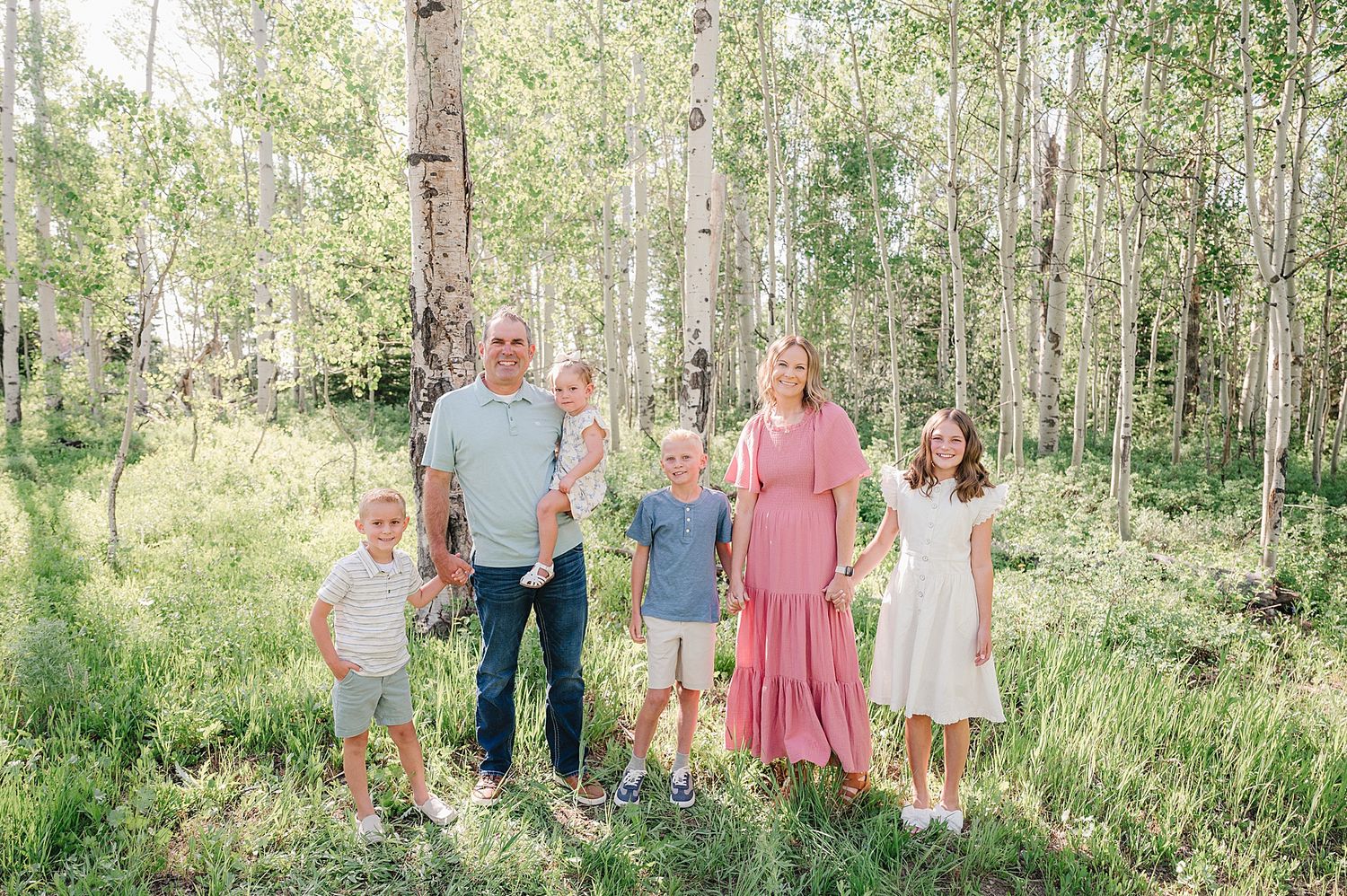 A family stands together in a sunlit aspen grove wearing coordinated pink and blue outfits for a summer photo session.
