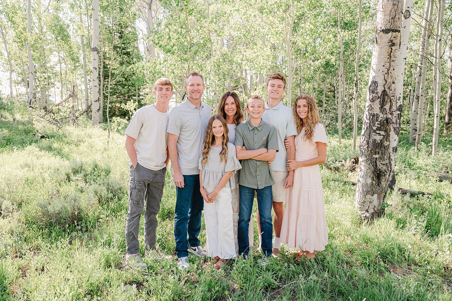A family group stands together outdoors in a sunlit aspen grove during summer.