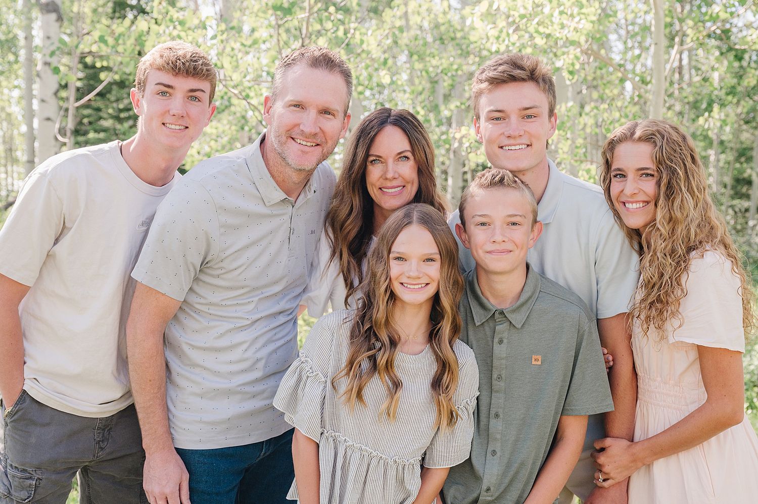A family poses together outdoors for a portrait in coordinated casual outfits with a natural forest background.