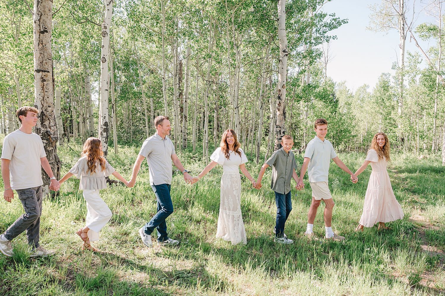 A group of people walking together through a sunlit aspen forest wearing light-colored casual clothing.