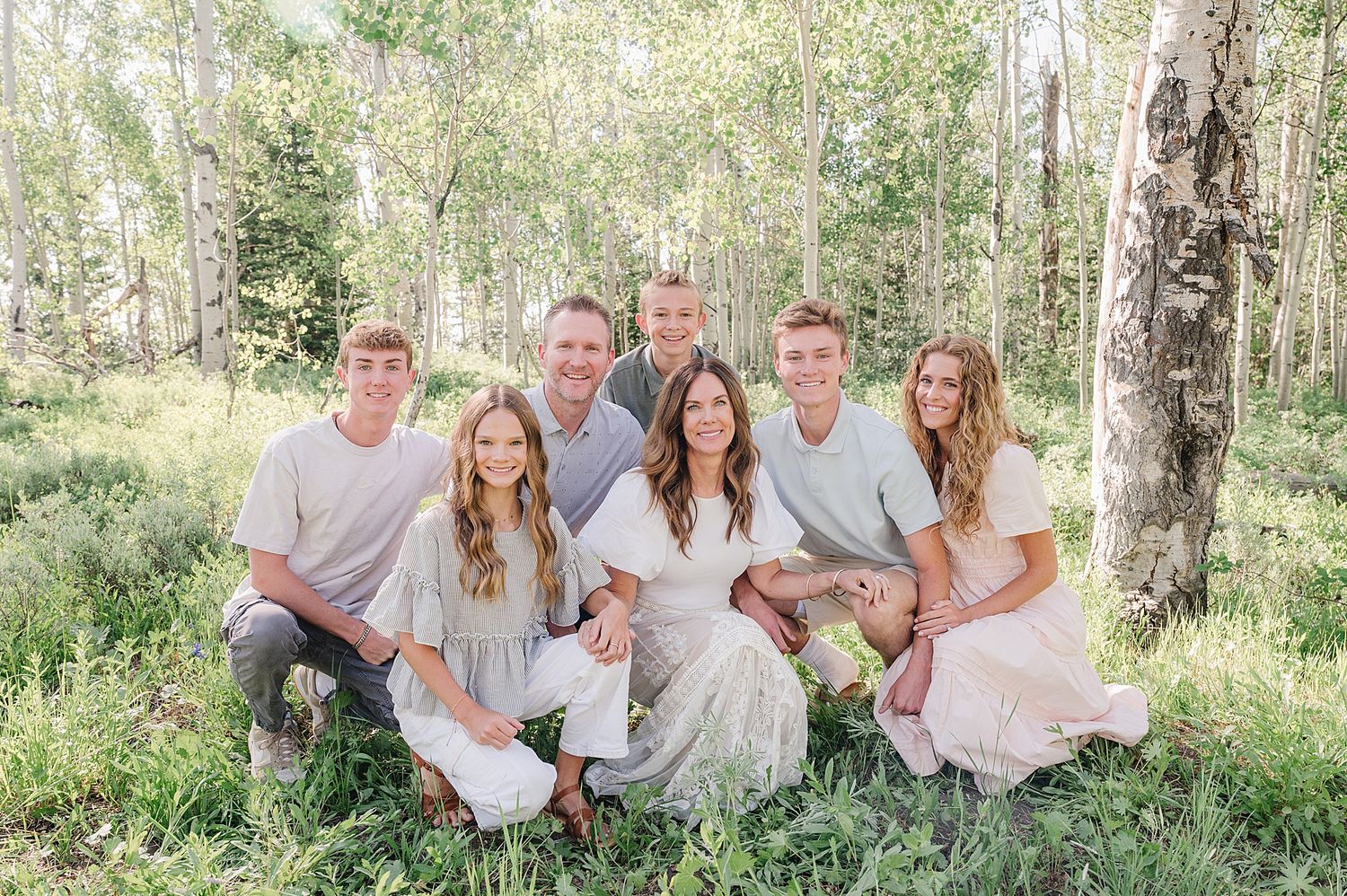 A group in coordinated light colored outfits sits together outdoors among aspen trees in a sunny forest setting.