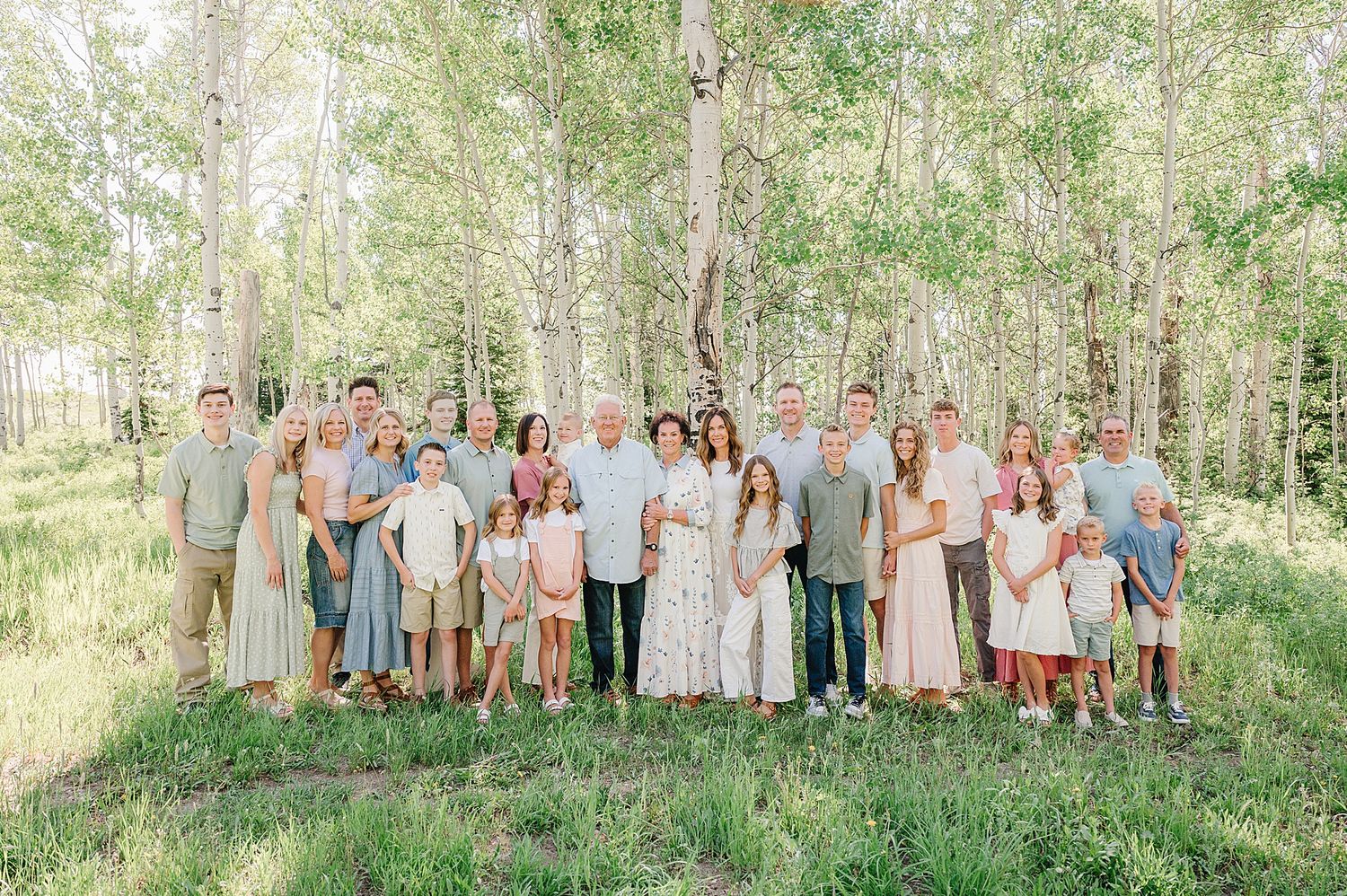 A large group in light-colored attire poses together for outdoor photography in a wooded area.