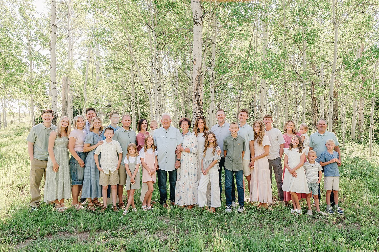 Large family gathering poses in a sun-dappled forest wearing coordinated pastel and neutral colored outfits.