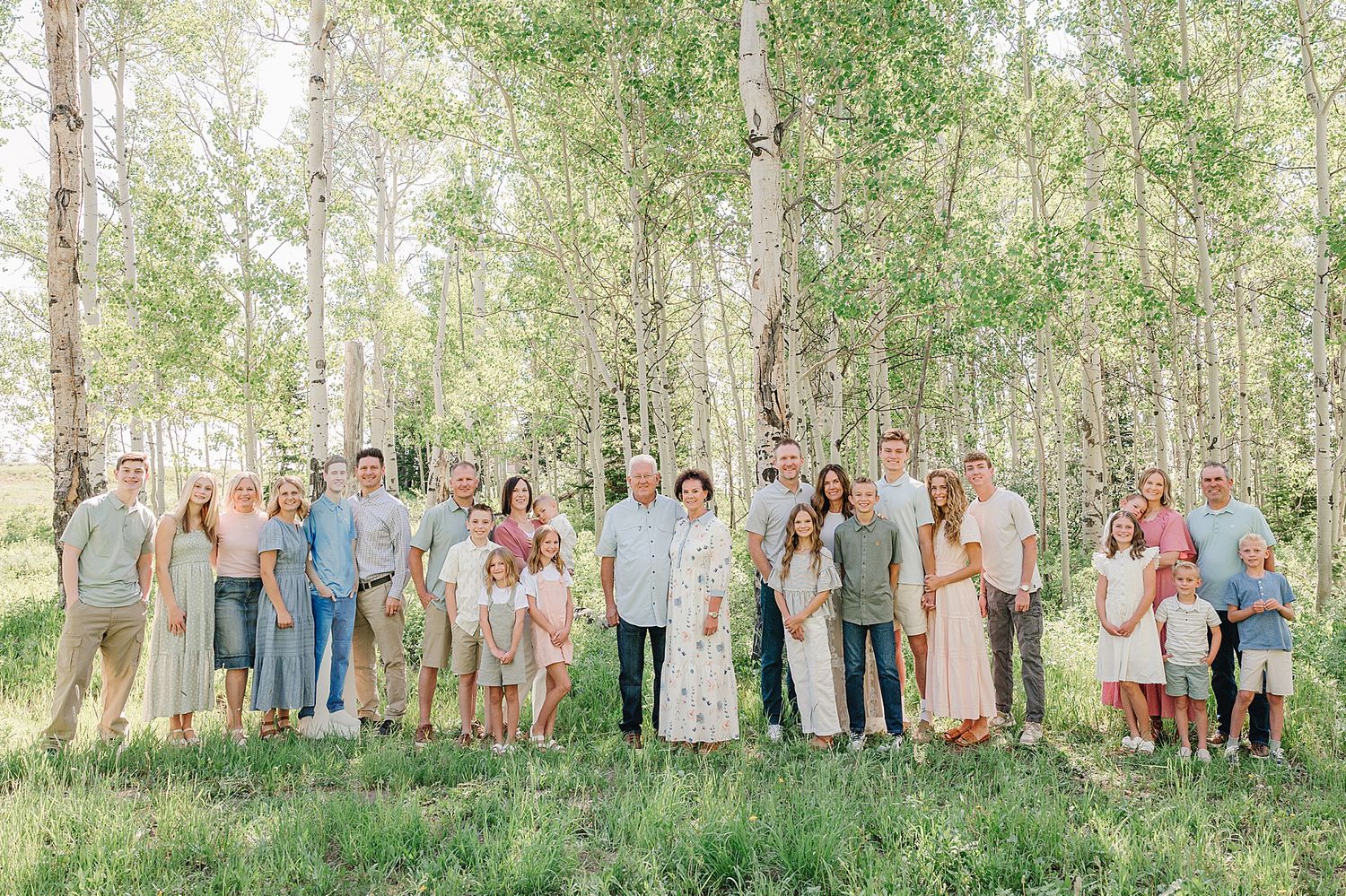 A large group stands together in casual attire among aspen trees in a sun-dappled forest setting.