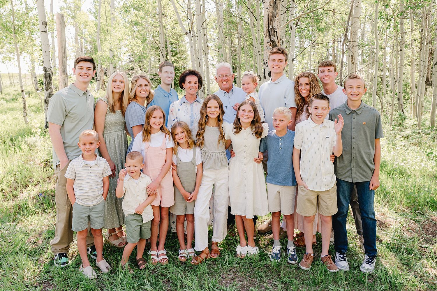 Extended family group dressed in coordinating neutral tones poses together outdoors in a sunlit wooded setting.