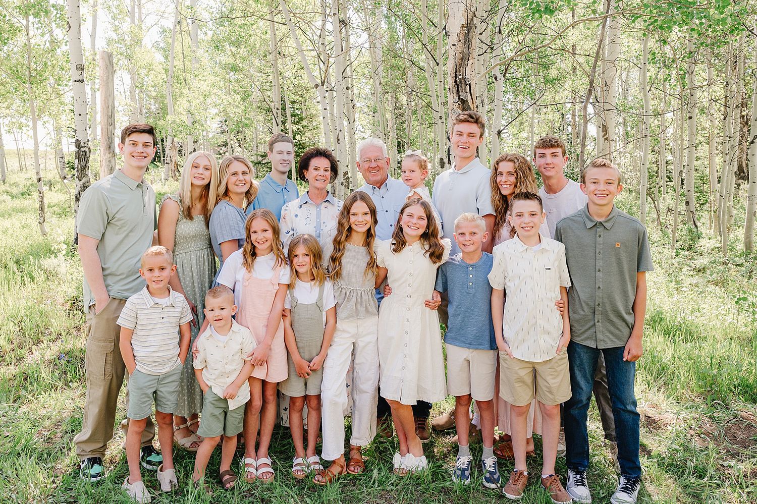 A large extended family group poses outdoors together in a wooded setting wearing coordinated casual summer clothing.