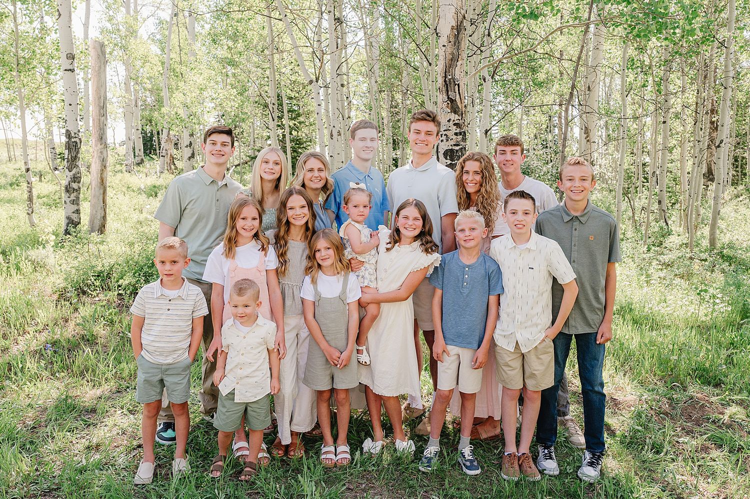 A large group of people wearing coordinated neutral-toned outfits poses together in a sunny forest setting.