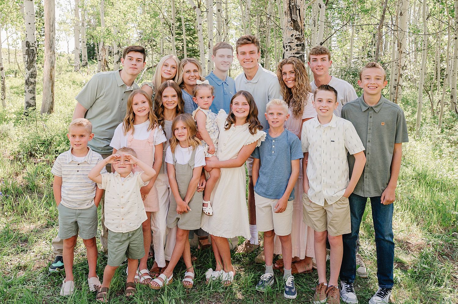 A large group wearing coordinated casual outfits poses together in a sunlit wooded outdoor setting.