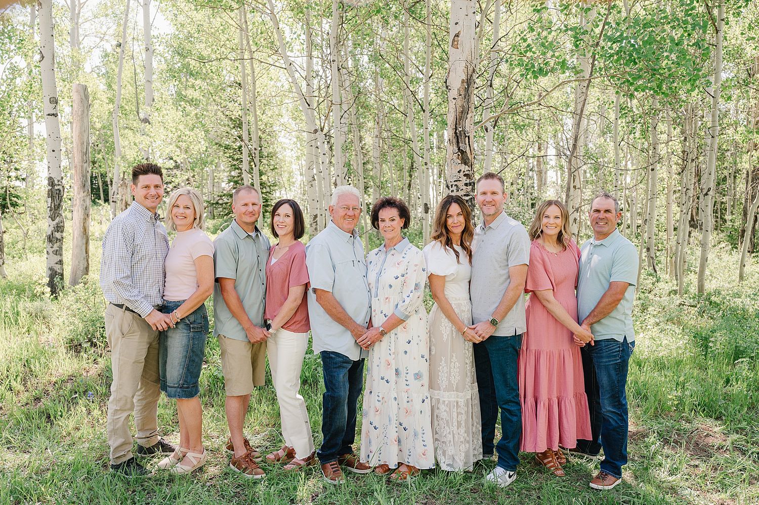 A group of people in coordinated pastel outfits pose together for a portrait in a sunlit wooded area.