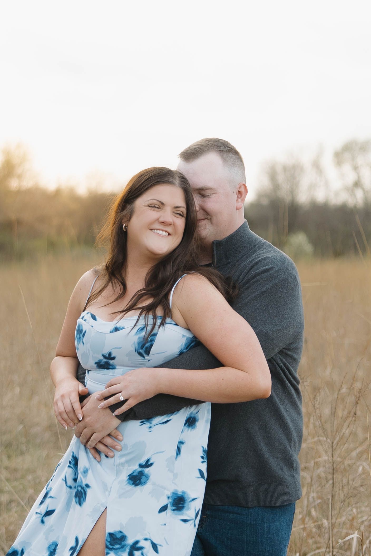 A romantic couple embraces in a golden field at sunset, with the woman wearing a blue floral dress.