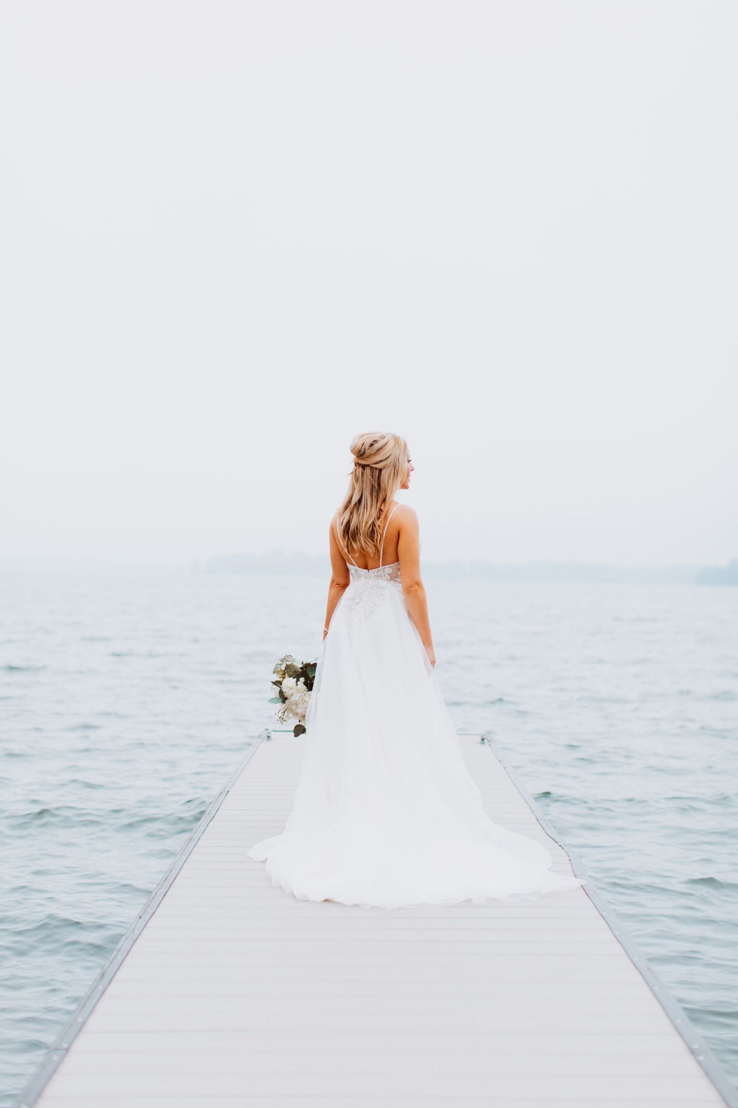 A bride in a flowing white gown stands on a wooden dock overlooking a misty ocean during golden hour.