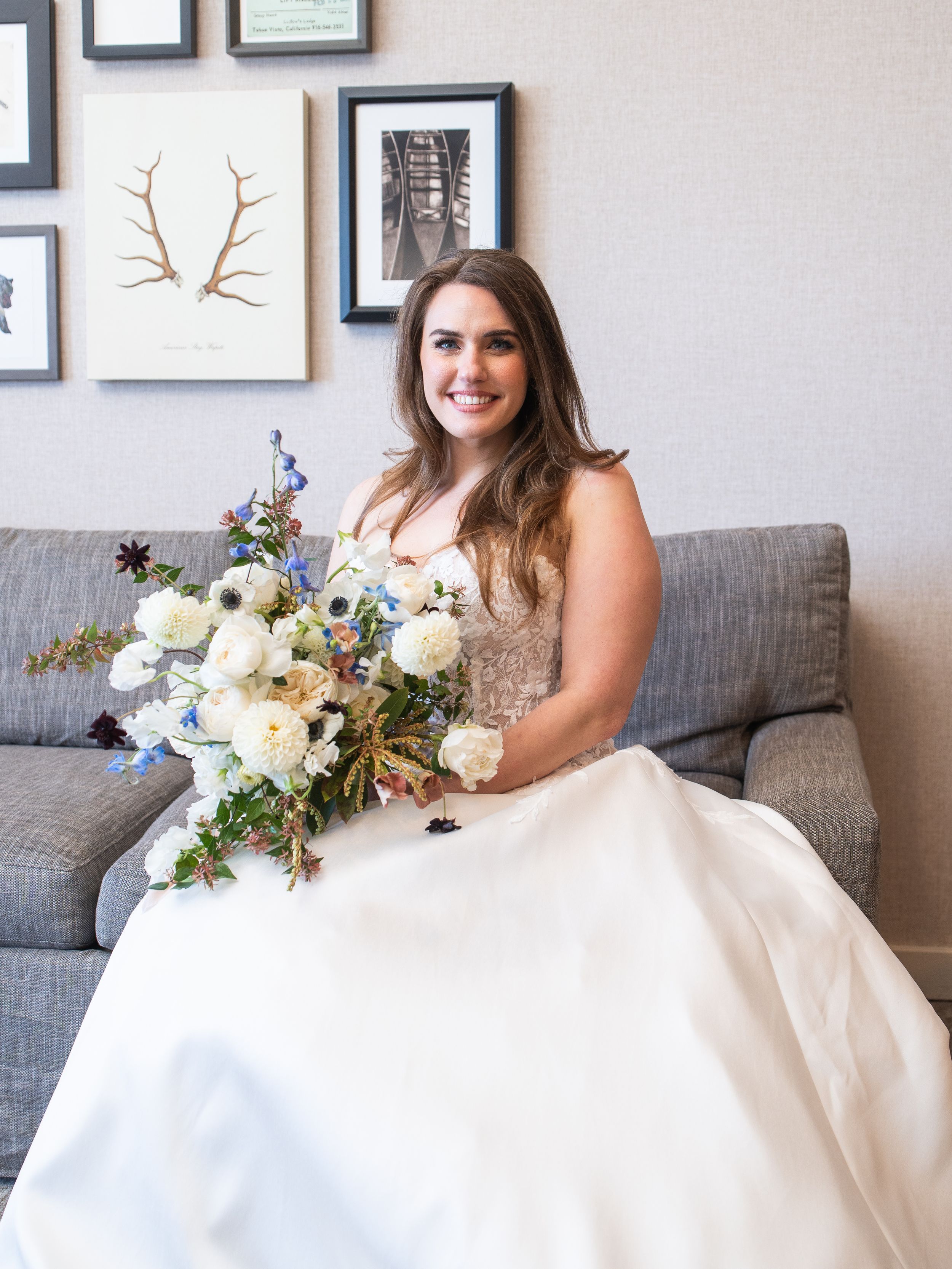 A wedding bouquet rests in the lap of someone wearing a white gown while sitting on a grey couch beneath framed wall art.