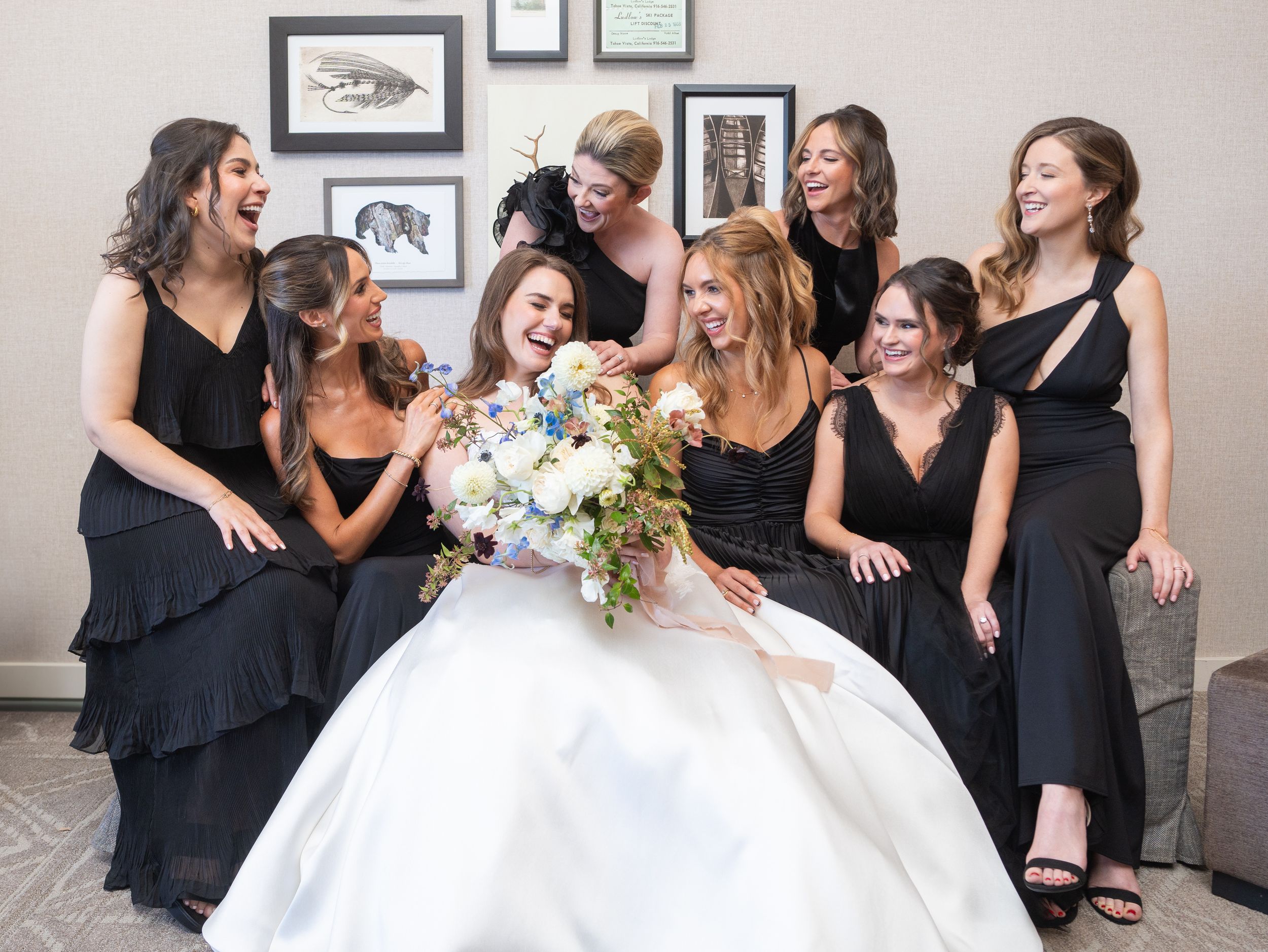 A bride in a white gown sitting with bridesmaids in black dresses share joyful laughter during a wedding celebration.
