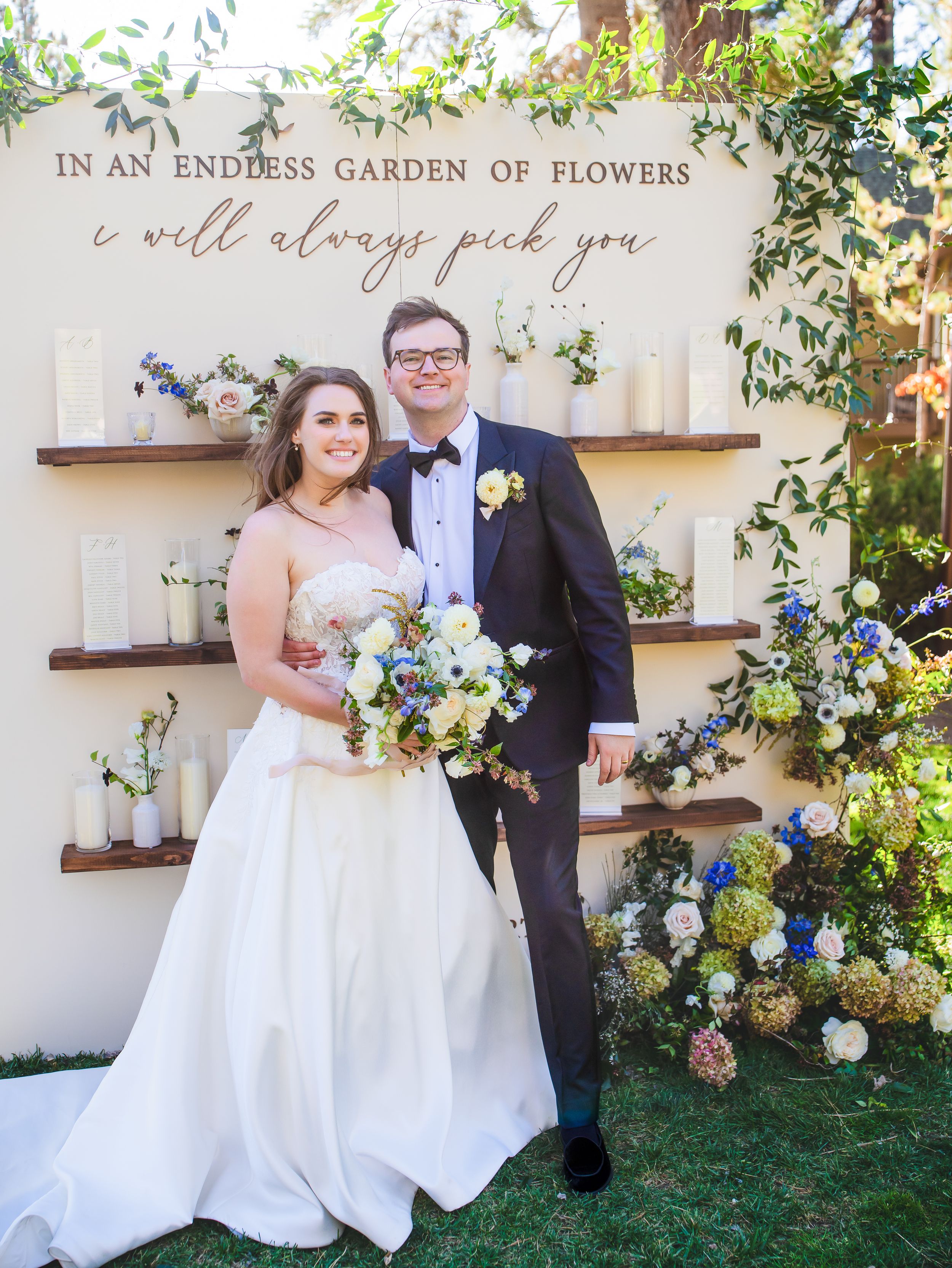 Wedding celebration scene with elegant floral wall display featuring 'endless garden of flowers' text and rustic wooden shelving.