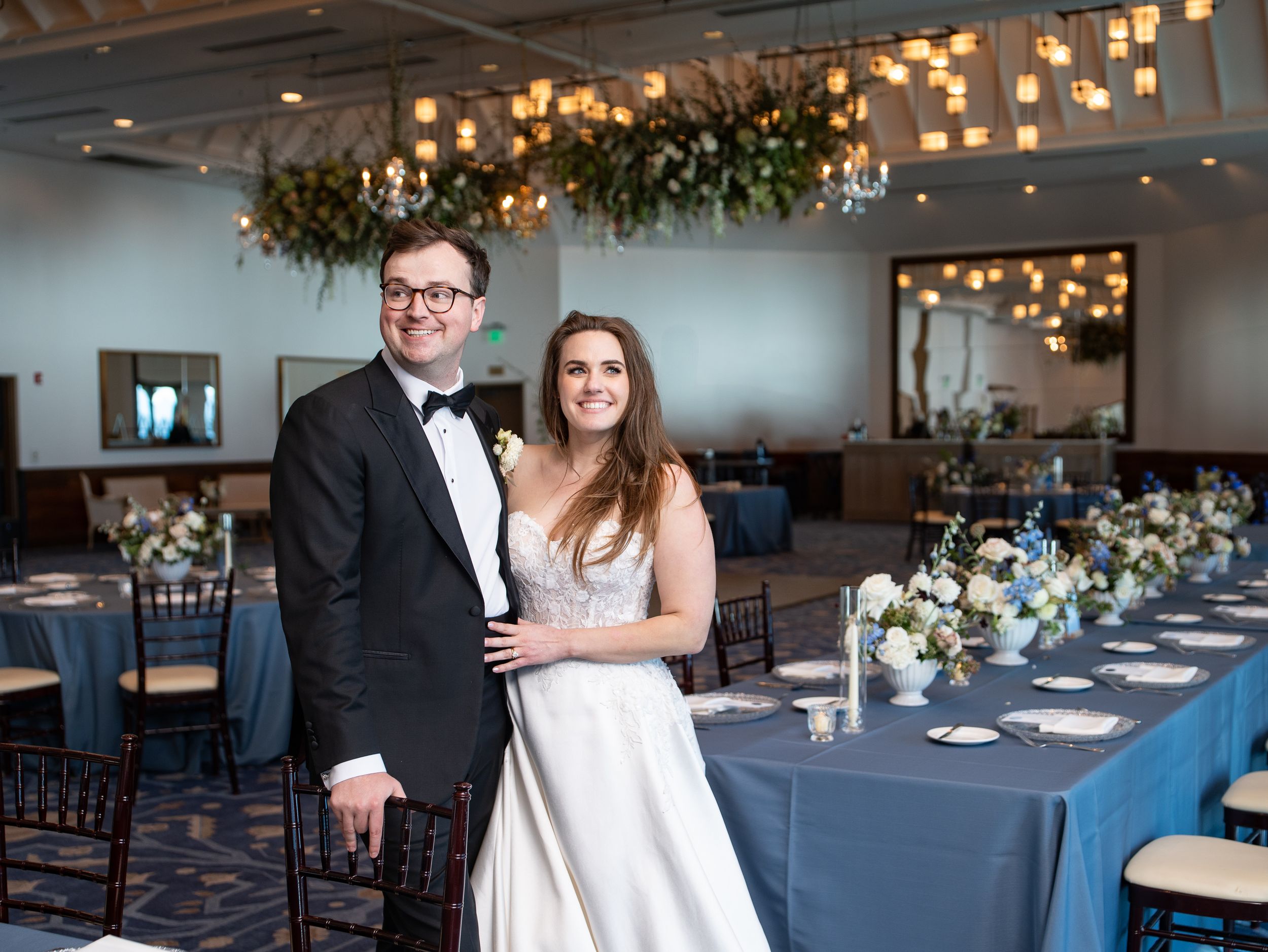 A couple poses in formal wedding attire inside an elegant reception venue with blue tablecloths and hanging floral decor.
